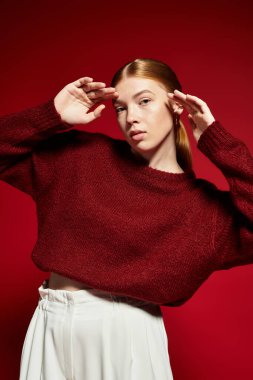 Şık bir kış süveteri giymiş kızıl saçlı bir kadın cesur bir kırmızı zemine karşı poz veriyor..