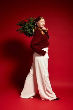 Sıcak süveter giyen neşeli genç bir kadın kırmızı bir fonda çam ağacını tutuyor..