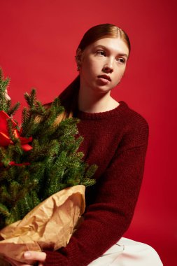 Samimi bir süveter giyen neşeli bir kızıl, küçük bir çam ağacıyla poz verir, bayram neşesini yayar..