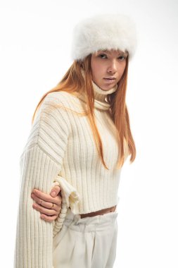 Genç bir kızıl şık bir şekilde rahat bir kazak ve kürk şapkayla poz veriyor. Kış havası için mükemmel..