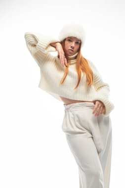 Genç kızıl saçlı bir kadın, hoş beyaz bir kazak ve kürk şapkayla, zarif bir şekilde poz vererek kış cazibesini sergiliyor..