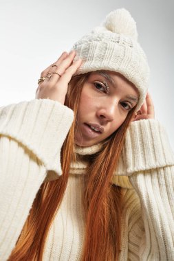 Genç bir kızıl saçlı şık bir şapka ve kalın bir süveter giymiş beyaz bir fona karşı poz veriyor..