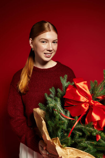 Веселая рыжая женщина в уютном свитере позирует с праздничным деревом и яркой лентой.