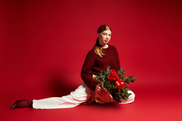 A young redhead poses gracefully in a winter sweater, holding a pine tree against a red backdrop.