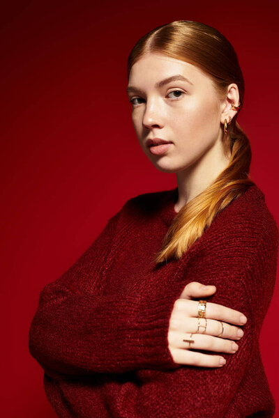 A young woman with red hair poses in a cozy sweater against a bright red background.
