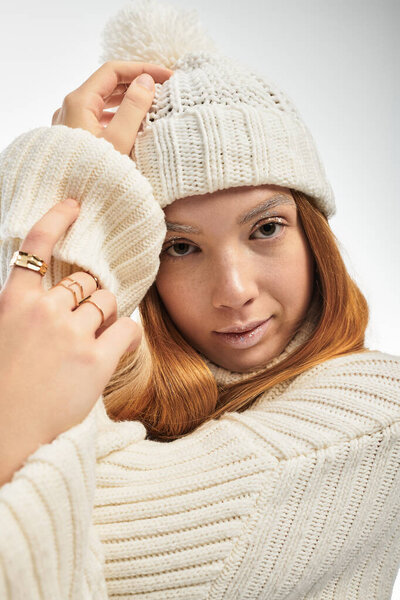A young woman with red hair poses against a white background, wearing a hat and knitted sweater.