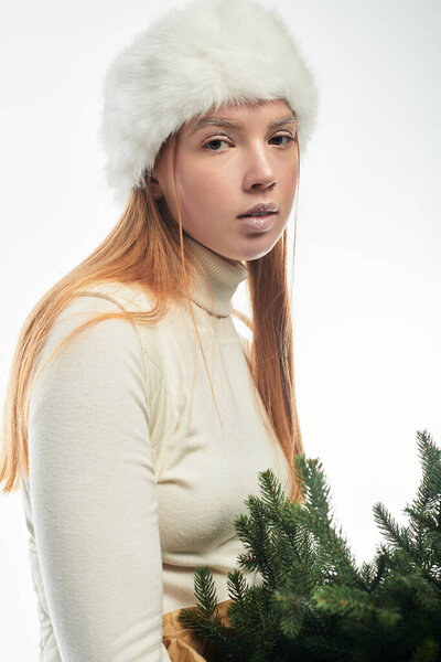 A young redhead poses in a soft sweater and fur hat, holding a small Christmas tree.