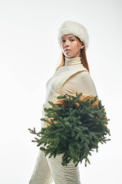 Redhead woman poses warmly in a cozy sweater and fur hat, holding a small Christmas tree.