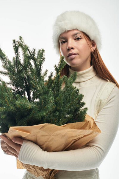 A young woman with red hair smiles while holding a small Christmas tree.