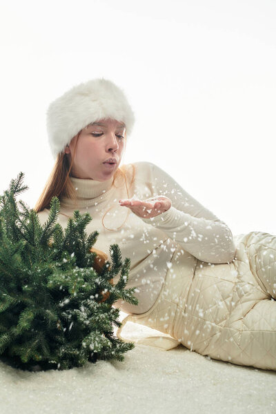 Молодая рыжая в меховой шляпе дует снежинками в окружении маленькой рождественской елки.