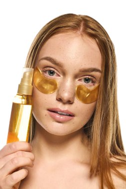 Genç bir kadın serumu tutarken altın göz bandı yapıştırıyor. Rahatlamış bir benlik bakımı anı sergiliyor..