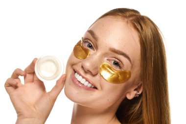 Parlak, neşeli genç bir kadın altın maskeler takarken elinde bir kavanoz cilt ürünü tutuyor..