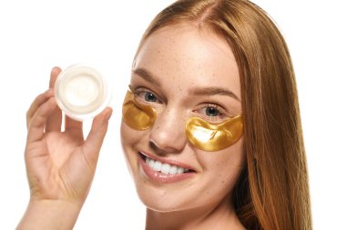 Altın göz bantları olan genç bir kadın elinde bir kavanoz cilt bakımıyla gülümsüyor..