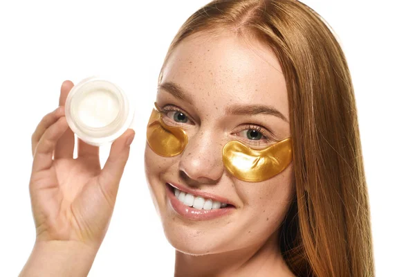 Altın göz bantları olan genç bir kadın elinde bir kavanoz cilt bakımıyla gülümsüyor..