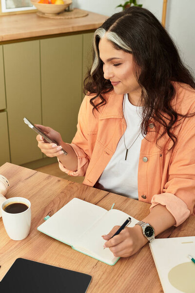 A woman with a white forelock sits at her table, focused on her phone and notebook.