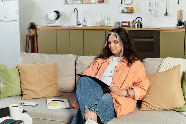 A woman with a white forelock relaxes at home, engaging with her notebook in a stylish apartment.