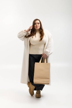 Samimi örgü süveter giyen genç bir kadın, parlak bir stüdyoda elinde bir alışveriş çantasıyla rahatça poz veriyor..
