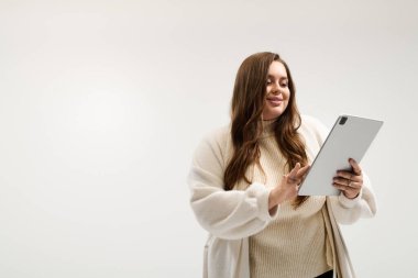 Kış süveteri giymiş iri cüsseli bir kadın parlak bir stüdyoda tabletle etkileşime geçiyor..