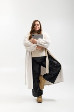 Geniş bedenli genç bir kadın, parlak bir stüdyoda örülmüş bir kazakla güven içinde poz verir..