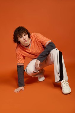 Kıvırcık saçlı genç bir çocuk günlük kıyafetlerle poz veriyor, modasını turuncuya karşı öne çıkarıyor..