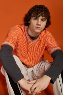 Kıvırcık saçlı genç bir çocuk, parlak turuncu gömlek ve pantolon giymiş, gelişigüzel oturuyor.