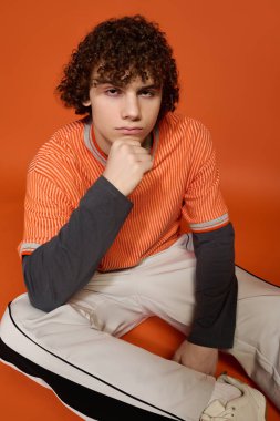 Kıvırcık saçlı bir genç, canlı turuncu bir arkaplana karşı düşünceli bir şekilde oturuyor ve tarzını sergiliyor..