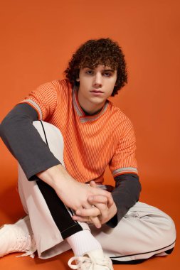 Kıvırcık saçlı genç bir çocuk turuncu bir arka planda günlük kıyafetlerle rahatlıyor..
