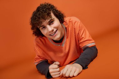 Rahat elbiseli, kıvırcık saçlı neşeli bir çocuk turuncu bir arka planda duruyor..