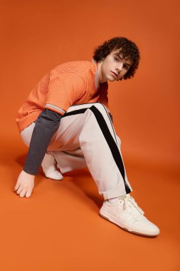 Kıvırcık saçlı bir çocuk öne eğiliyor, parlak turuncu bir fonda sokak tarzıyla hava atıyor..