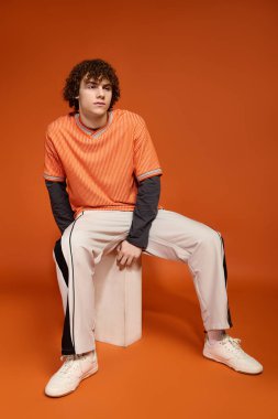 Kıvırcık saçlı, günlük kıyafetleri olan bir çocuk turuncu bir arka planda rahatça oturur..