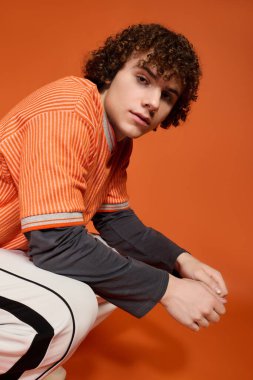 Kıvırcık saçlı bir genç, parlak turuncu bir arkaplanda şık bir şekilde oturur..