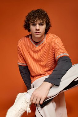 Kıvırcık saçlı genç bir çocuk turuncu bir arka planda şık kıyafetini sergiliyor..