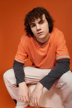 Kıvırcık saçlı genç bir çocuk turuncu arka planda gelişigüzel, sokak modasını sergiliyor..