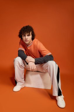 Kıvırcık saçlı genç bir çocuk, günlük modasını turuncu bir arka planda sergiliyor..