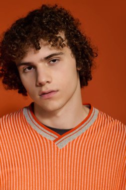 Gündelik giyinmiş genç bir kıvırcık çocuk parlak turuncu bir arka planla rahatça poz verir..