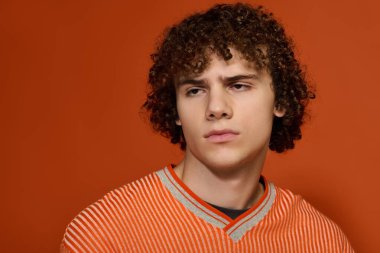 Kıvırcık saçlı genç bir çocuk parlak turuncu bir arka planda günlük kıyafetlerle poz veriyor..