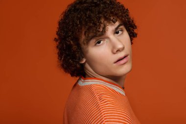 Kıvırcık saçlı genç bir çocuk turuncu bir arkaplanda rahat bir şekilde poz veriyor..