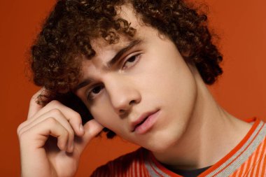 Kıvırcık saçlı genç bir çocuk parlak turuncu bir arka planda günlük giysiler içinde düşünceli poz verir..