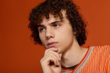 Genç, kıvırcık saçlı bir çocuk parlak turuncu bir arka planda düşünceli bir şekilde poz verir..