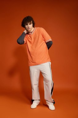 Kıvırcık saçlı genç bir çocuk kendine güveniyor, parlak turuncu bir arka planda günlük kıyafetler giyiyor..