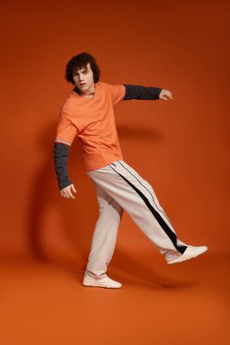 Kıvırcık saçlı genç bir çocuk parlak turuncu bir arka planda günlük giysiler içinde, enerji saçıyor..