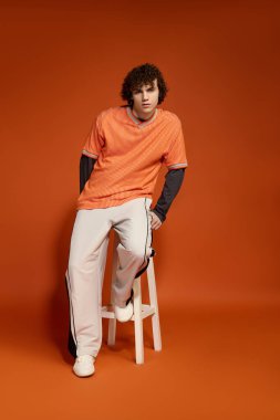 Günlük kıyafetli genç bir çocuk canlı turuncu ile çevrili bir taburede güven içinde poz verir..