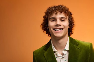 Kıvırcık saçlı genç bir çocuk şık yeşil kadife bir ceket giyerken ışıl ışıl gülümsüyor..