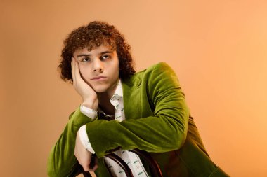 Kıvırcık saçlı genç bir çocuk bir stüdyoda yeşil kadife ceket giyerek çenesini uzatıyor..