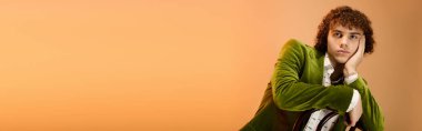 Kıvırcık bir genç, yeşil kadife bir ceketle düşünceli bir şekilde poz veriyor, turuncu bir fonda, pankartta.