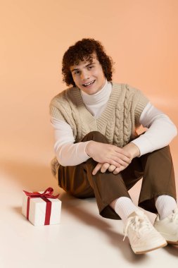 Kıvırcık bir genç çocuk stüdyo zemininde oturuyor ve Noel hediyesinin yanında gülümsüyor..