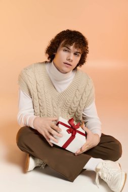 Kıvırcık saçlı genç elinde bir hediye kutusuyla stüdyoda oturuyor. Kış tatili kutlamalarına hazır..