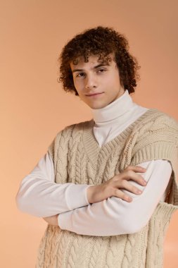 Şık kıvırcık saçlı bir genç örgü bir yelek giyer ve rahat bir stüdyo ortamında poz verir..