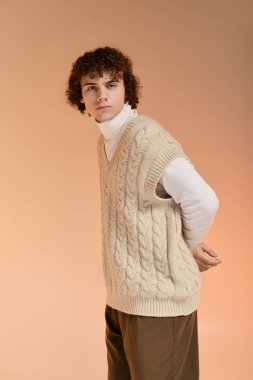 Kıvırcık saçlı genç bir çocuk stüdyo ortamında şık örülmüş bir yelek gösteriyor..