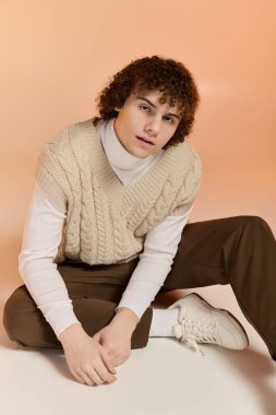 Kıvırcık saçlı genç çocuk yerde oturur ve kış için şık bir örgü yelek modeller..